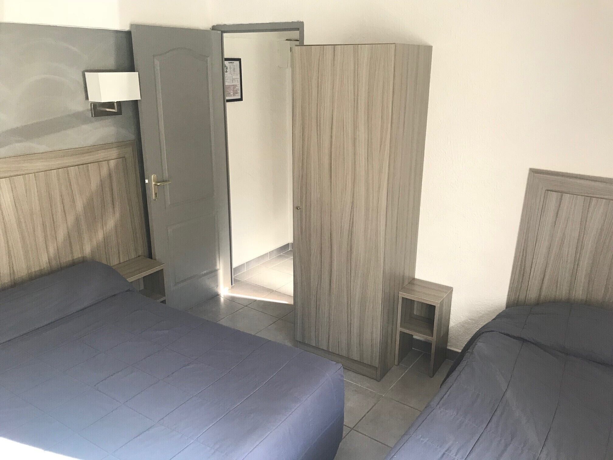 A hotel room with two beds, a wardrobe, a bedside table, and a wall-mounted lamp.