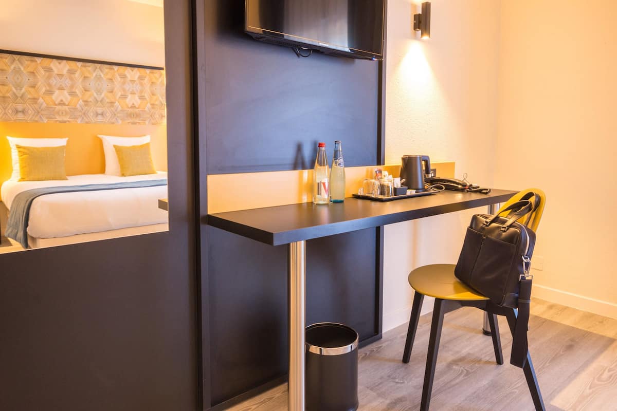 A hotel room with a bed, a desk with a chair, a TV, and a trash can.