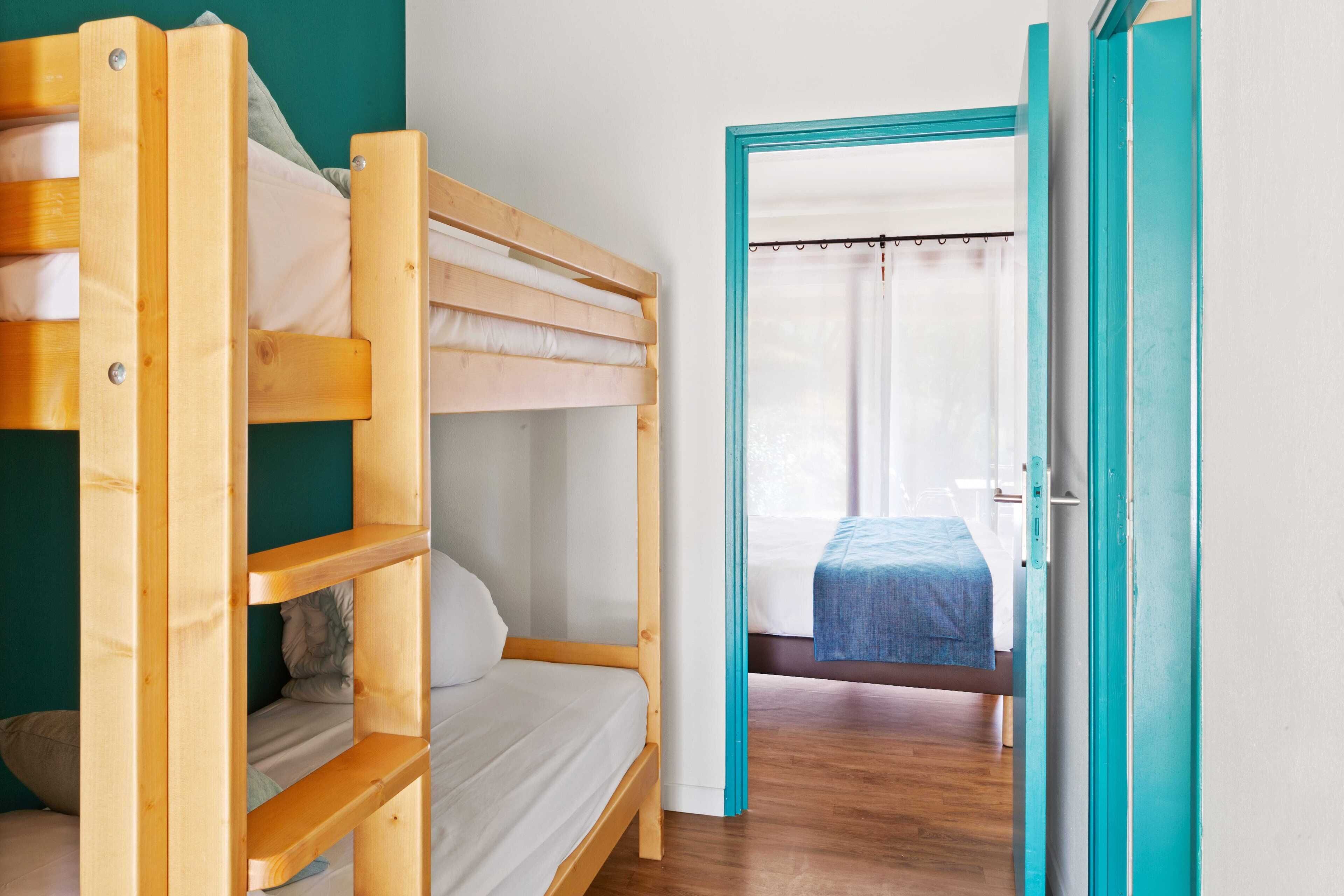 A bunk bed room with a wooden frame, a door leading to another room, and a visible bed with a blue blanket.