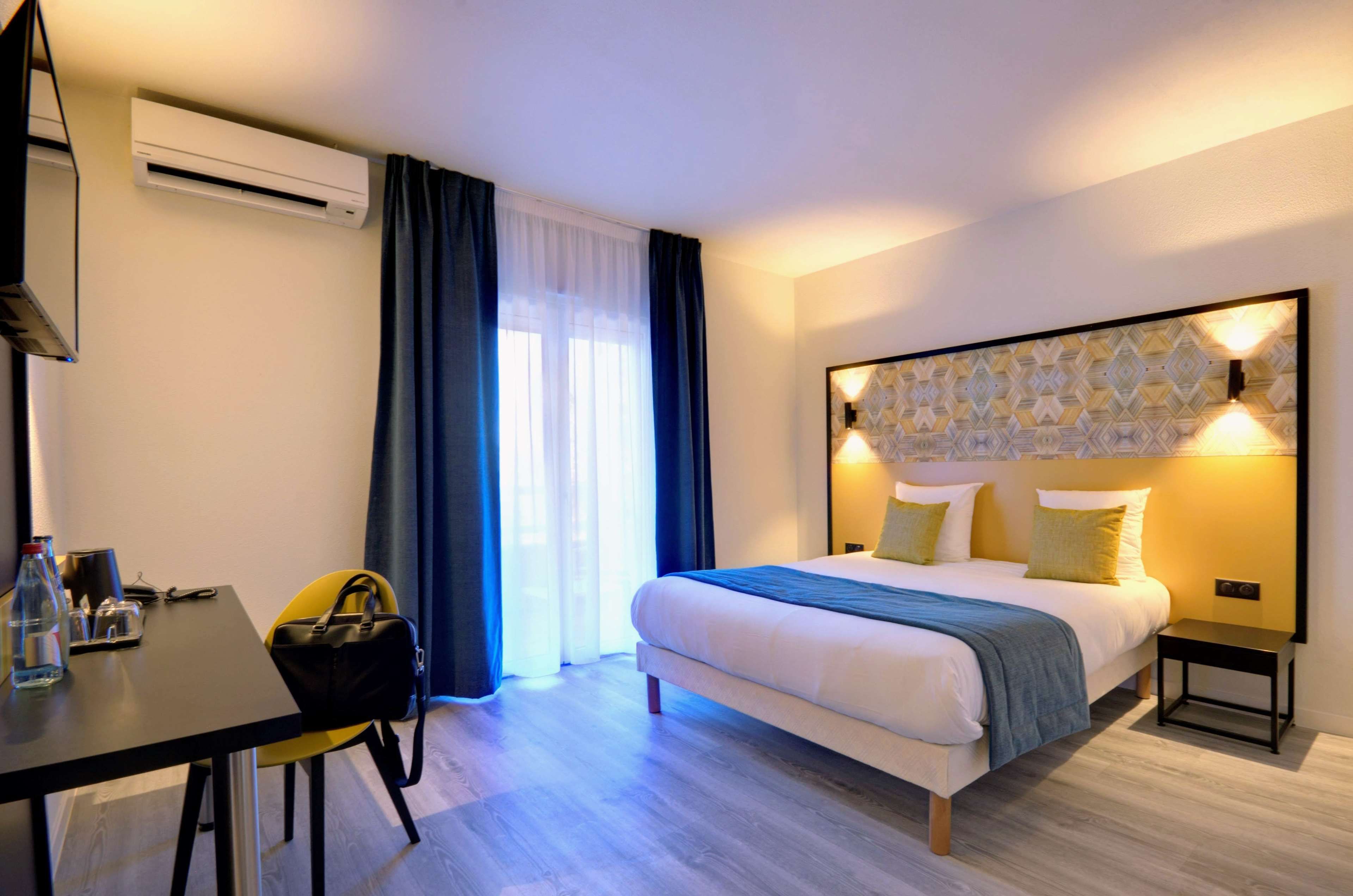 Una habitación de hotel con cama, mesita de noche, silla, escritorio y unidad de aire acondicionado.
