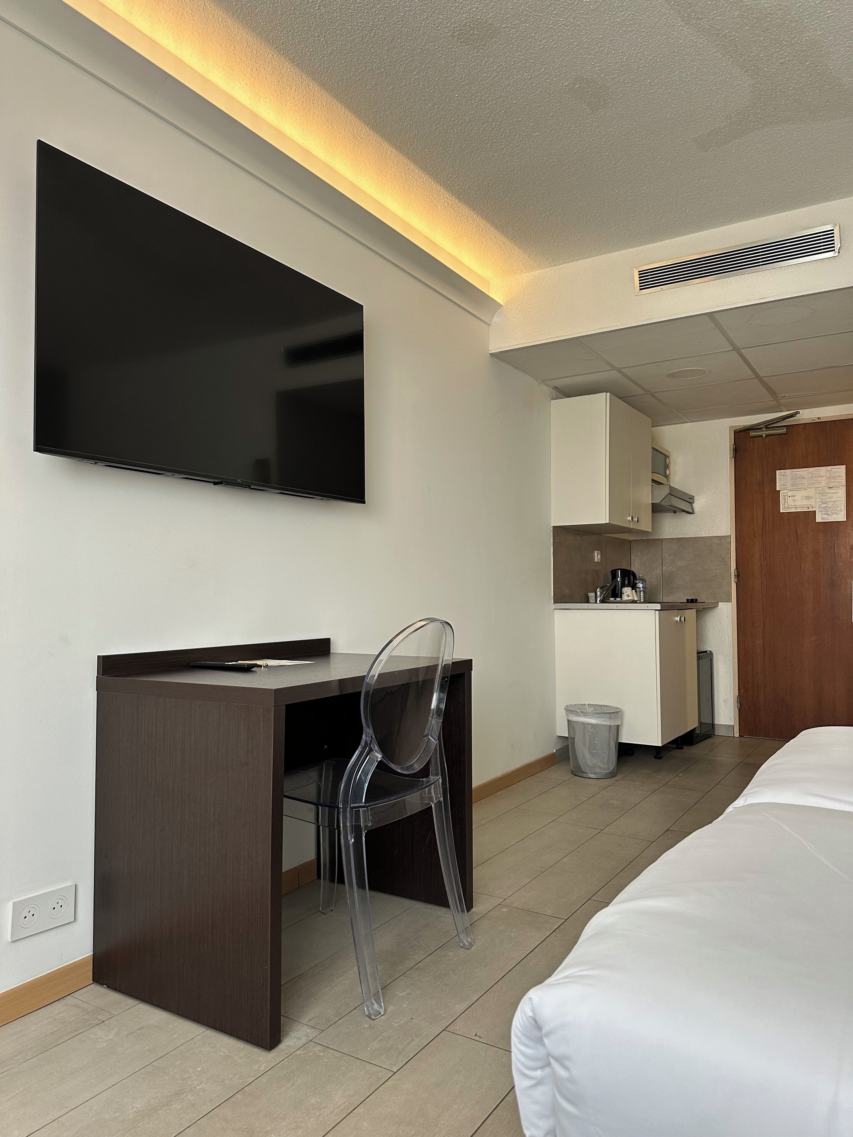A hotel room with a bed, a desk with a chair, a television, a kitchenette, and a door with a notice.