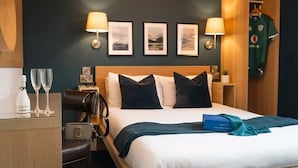 A hotel room with a bed, two bedside lamps, framed pictures on the wall, a minibar, a coat rack with a sports jersey, and a blue gift box. - Celtic Lodge Guesthouse - Restaurant & Bar (Dublin)