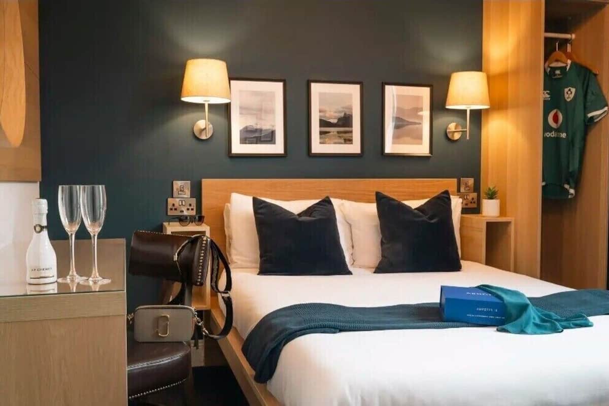 A hotel room with a bed, two bedside lamps, framed pictures on the wall, a minibar, a coat rack with a sports jersey, and a blue gift box.