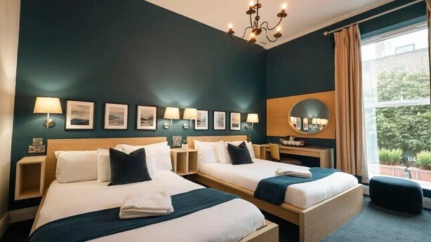 A hotel room with two beds, a chandelier, wall-mounted lamps, a mirror, and a window with a view of greenery. - Celtic Lodge Guesthouse - Restaurant & Bar (Dublin)
