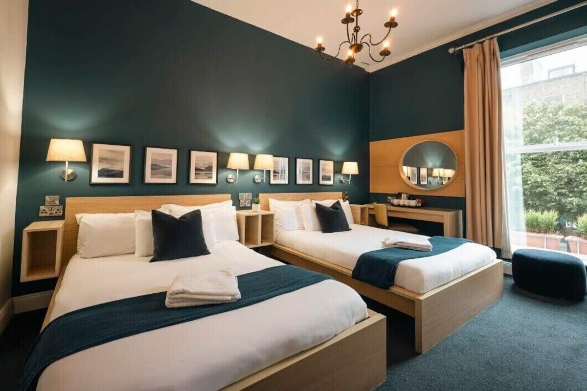 A hotel room with two beds, a chandelier, wall-mounted lamps, a mirror, and a window with a view of greenery.