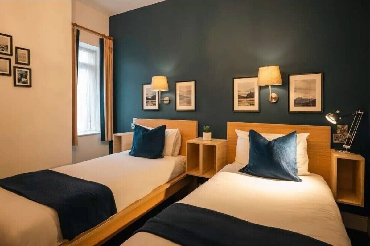 A hotel room with two single beds, each with a wooden headboard and a bedside table. There are wall-mounted lamps and framed pictures on the wall.
