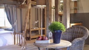 Una habitación rústica con vigas de madera, muebles de mimbre, una mesa redonda con una planta en maceta y una cama con cabecero blanco.