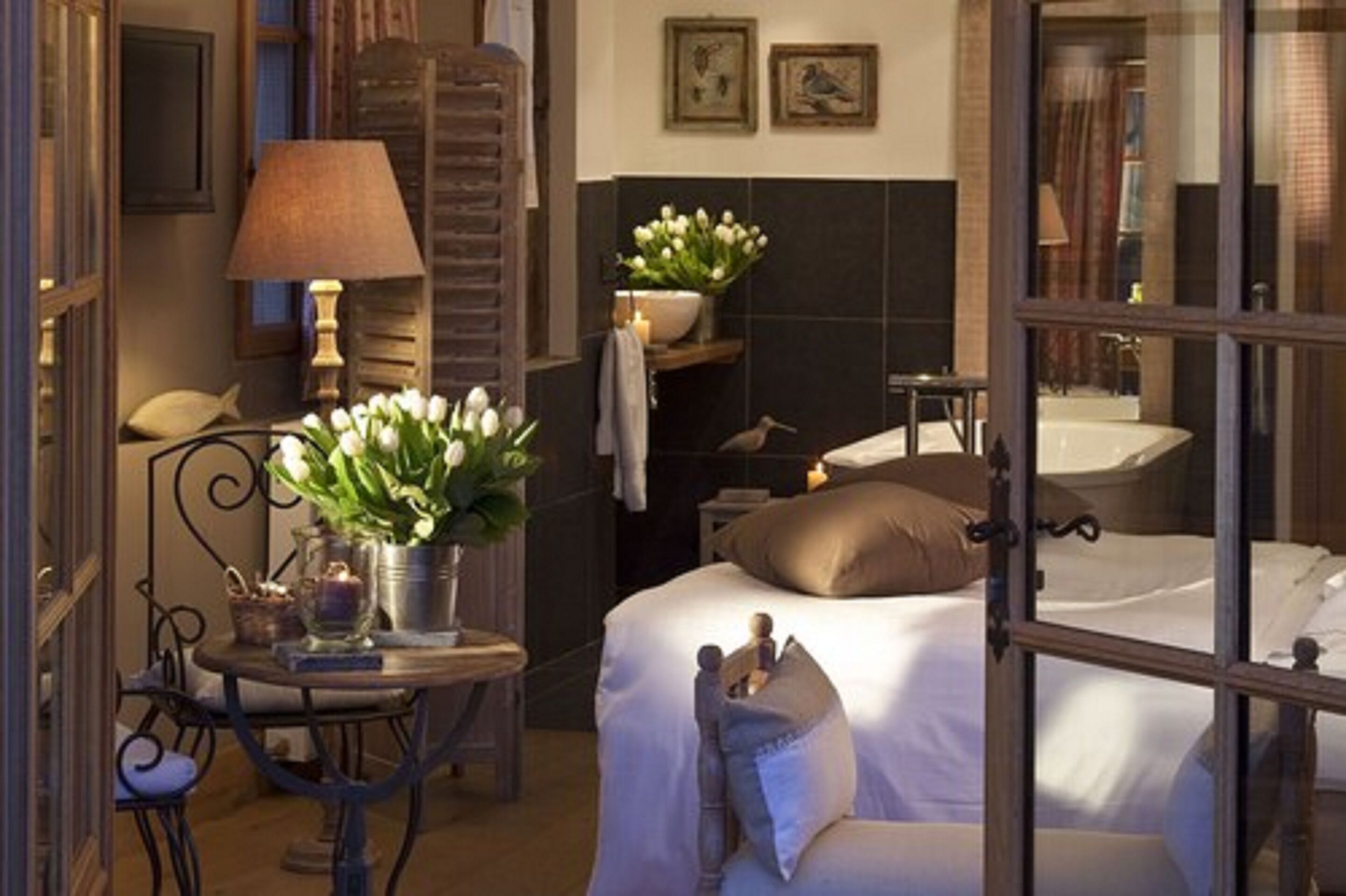 Habitación de hotel con una cama, una mesa con lámpara, un jarrón con flores y una ventana con vista al baño.