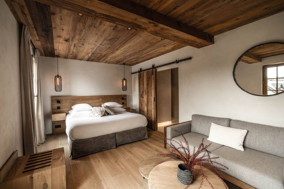 A modern bedroom with a wooden ceiling, a bed with a grey headboard, a grey sofa, and a round wooden table with a vase.