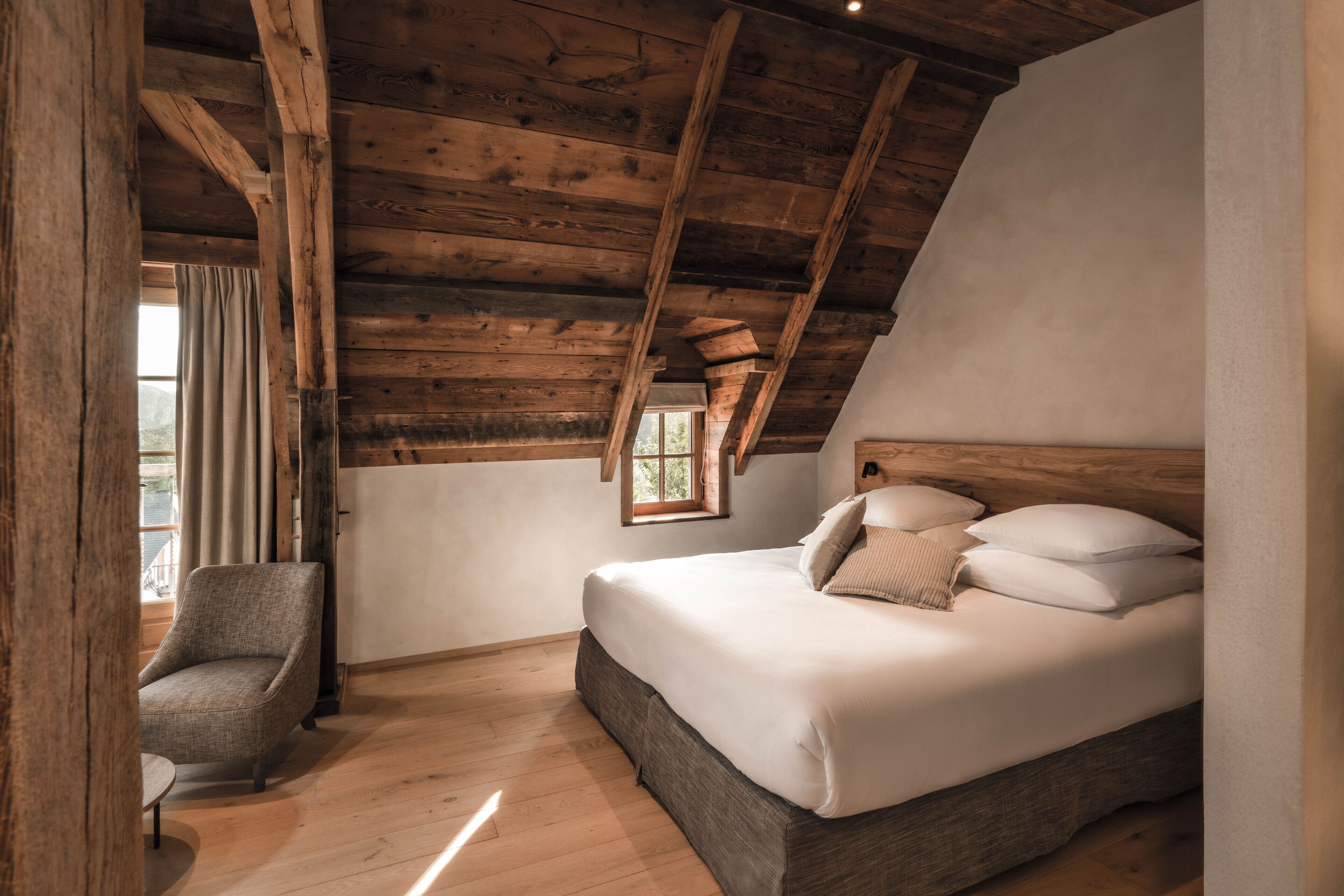 A bedroom with a wooden ceiling, a bed with white bedding, a chair, and a window.