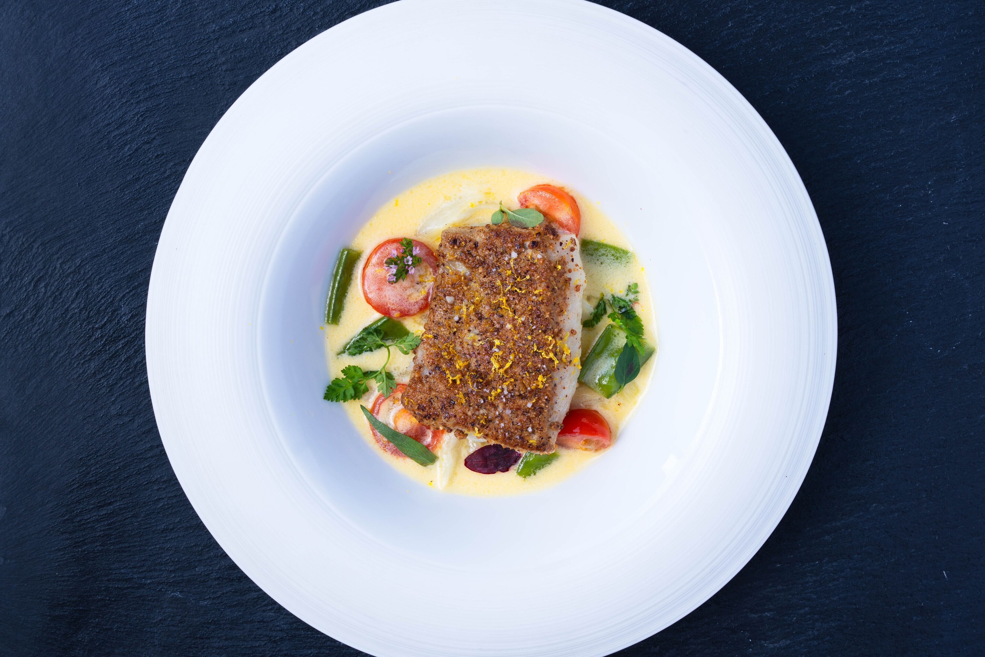 A white plate with a fish fillet, tomatoes, and green vegetables on a dark background.