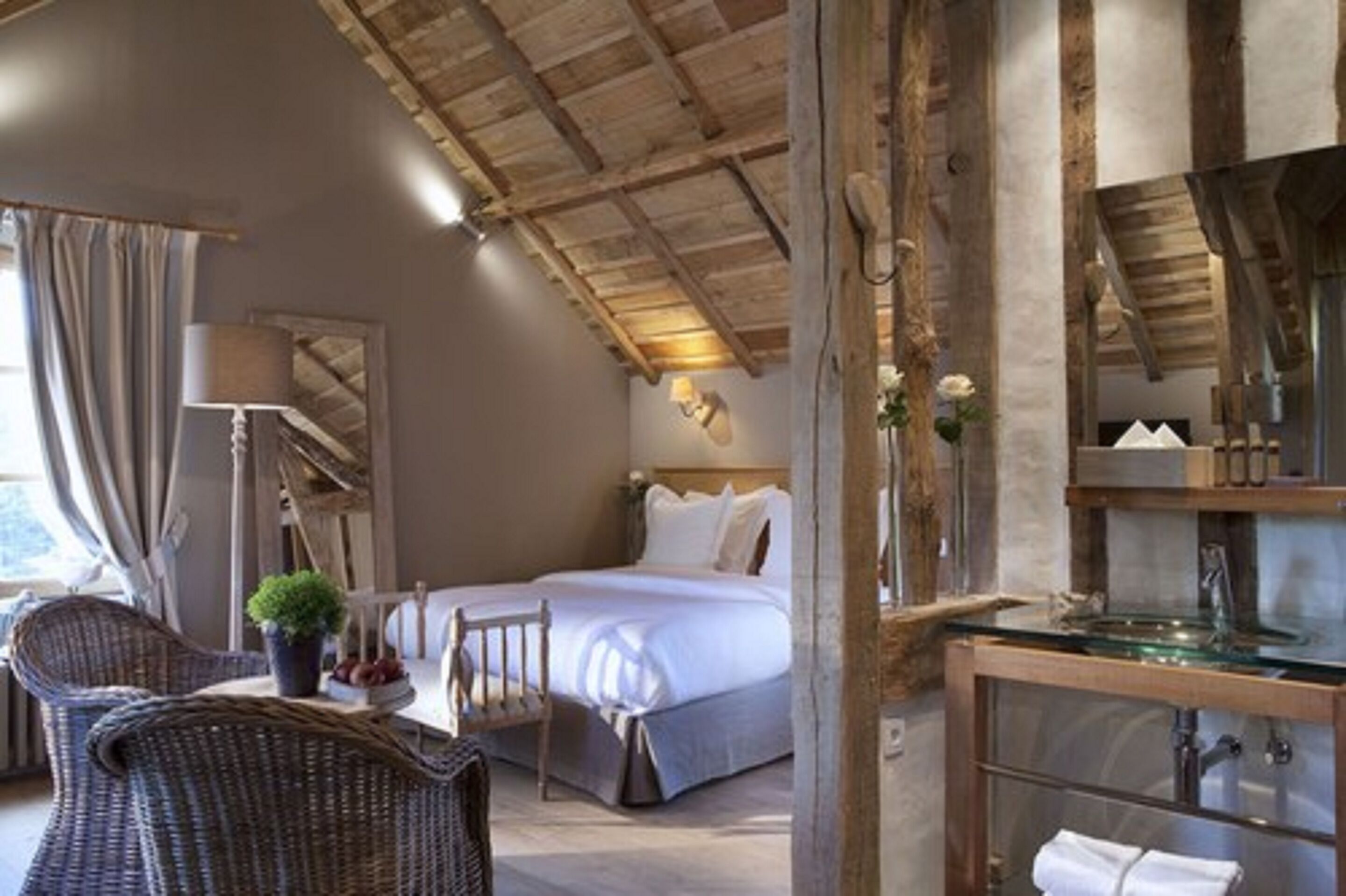 Ein Schlafzimmer mit einem Bett, einem Sessel, einem Tisch mit Pflanze, einer Lampe sowie einem Badezimmer mit Waschbecken aus Glas.