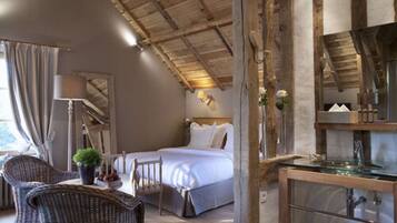 Ein Schlafzimmer mit einem Bett, einem Sessel, einem Tisch mit Pflanze, einer Lampe sowie einem Badezimmer mit Waschbecken aus Glas.