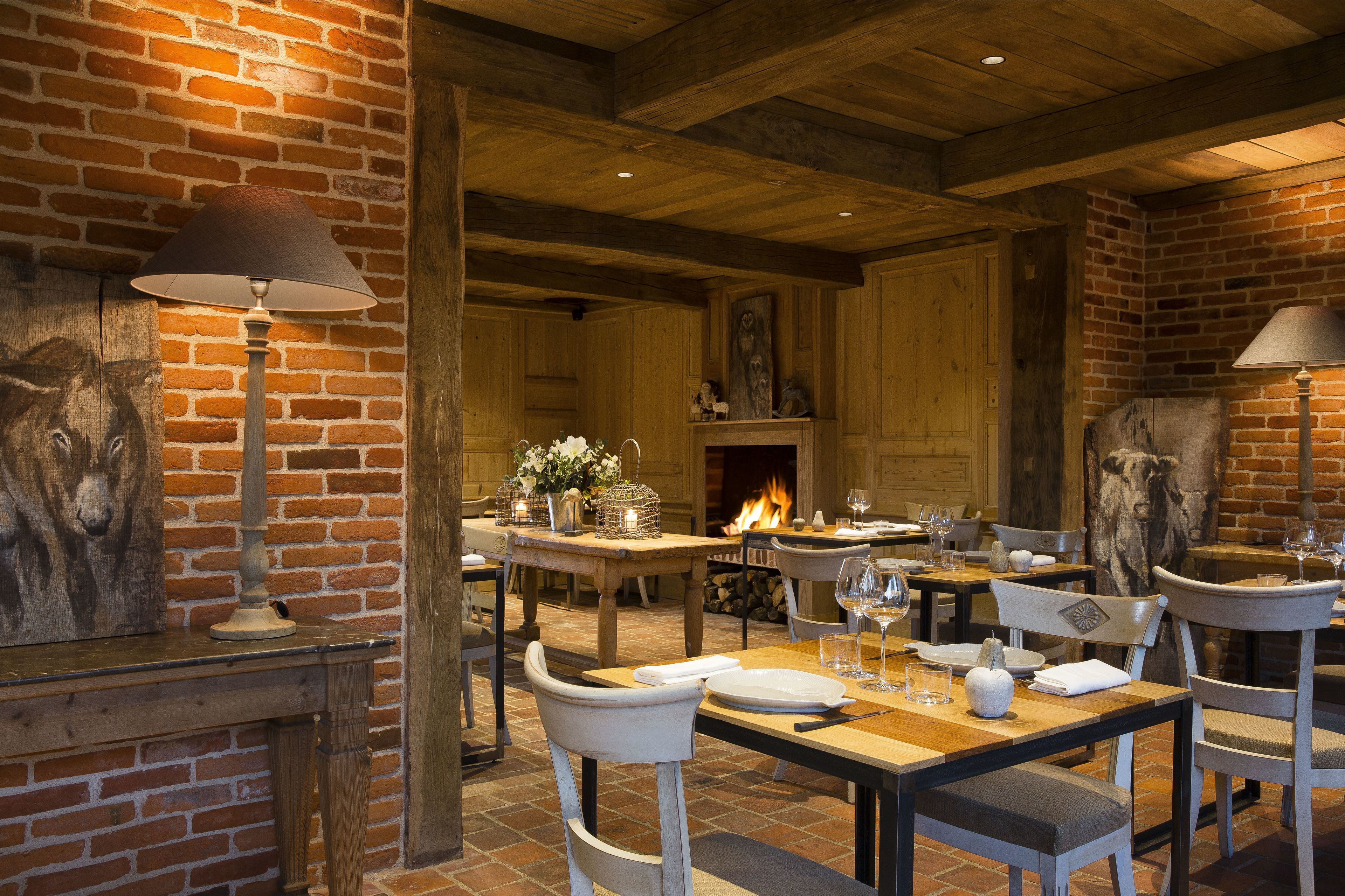 A rustic dining area with brick walls, wooden beams, and a fireplace. Tables are set with plates, glasses, and cutlery.
