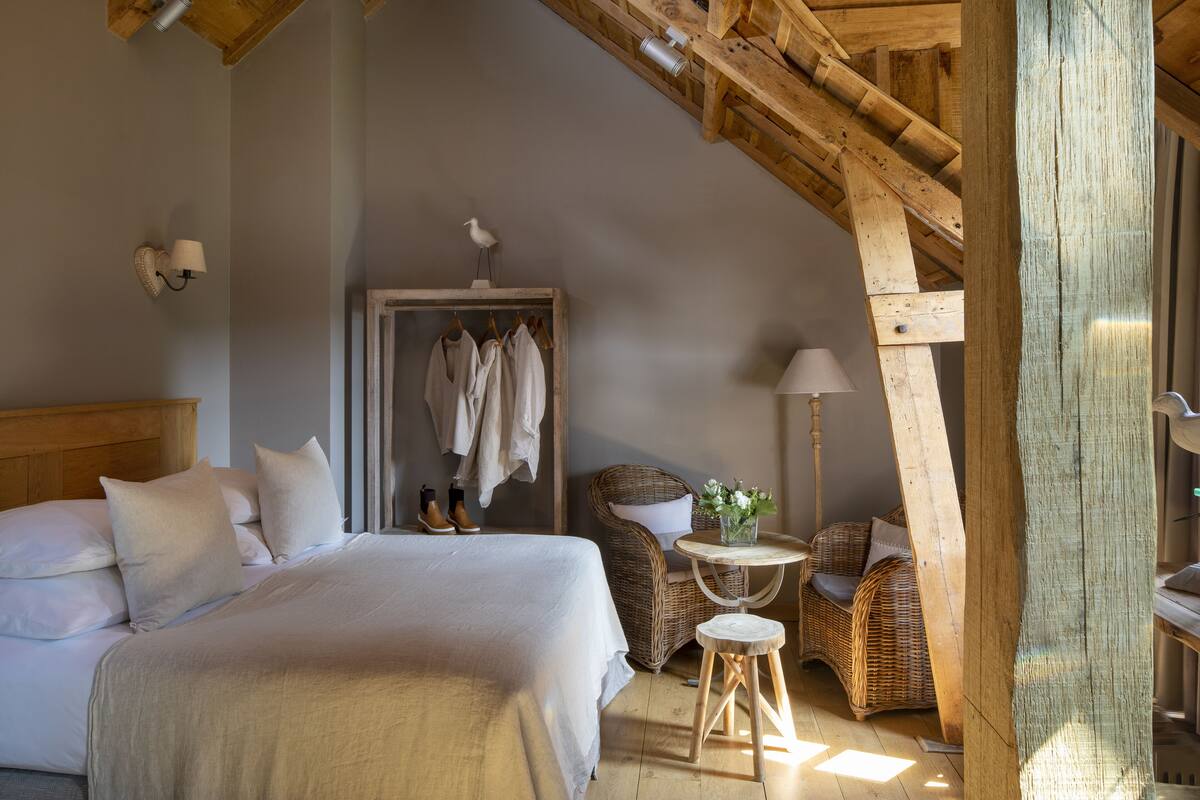A bedroom with a large bed, a wooden ceiling, a wicker chair, a small table, and a door with hanging clothes.