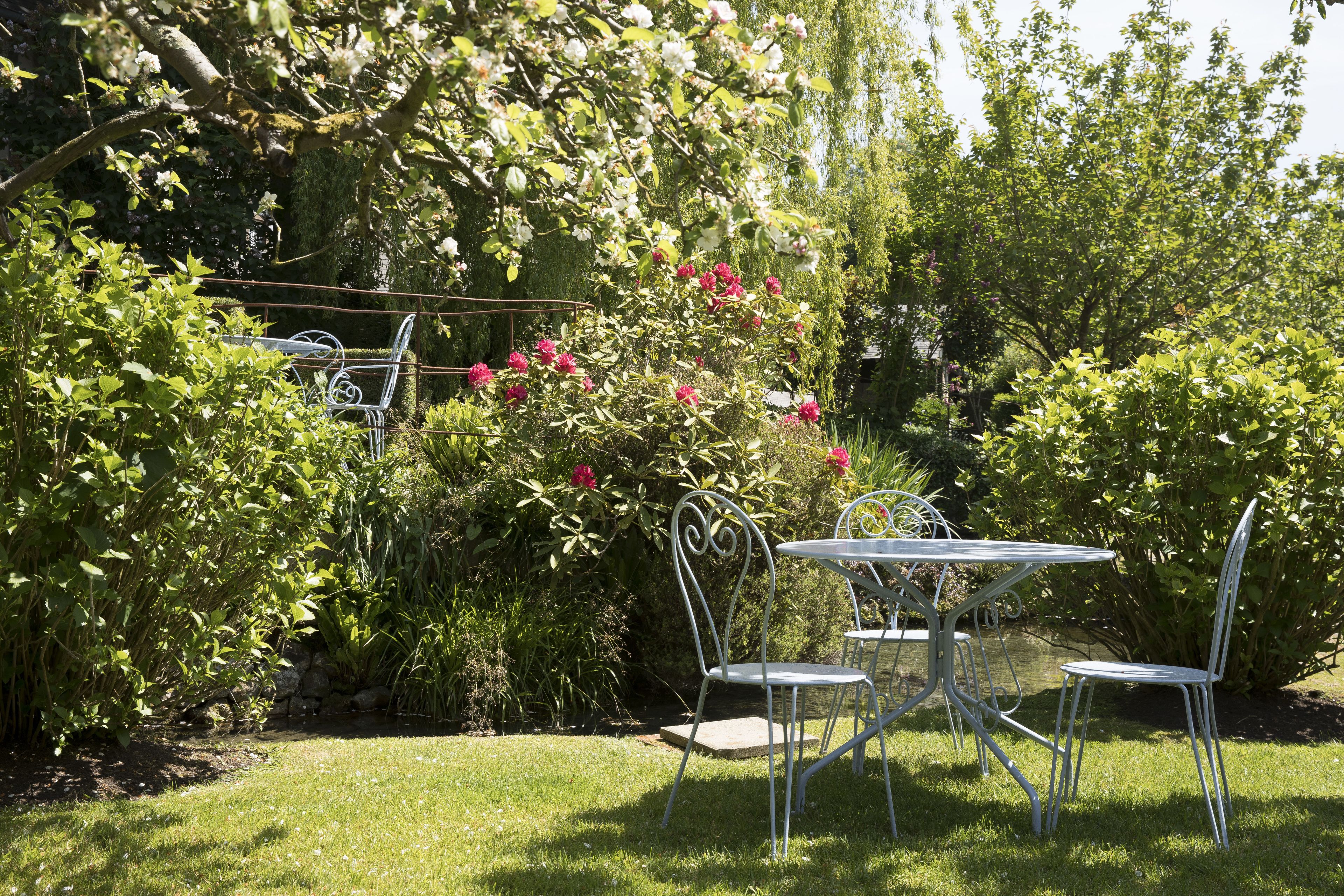 A garden with a metal table and chairs, surrounded by lush greenery and flowering plants.