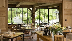 Un área de comedor al aire libre con mesas y sillas de madera, ventanas amplias y un techo rústico de madera.