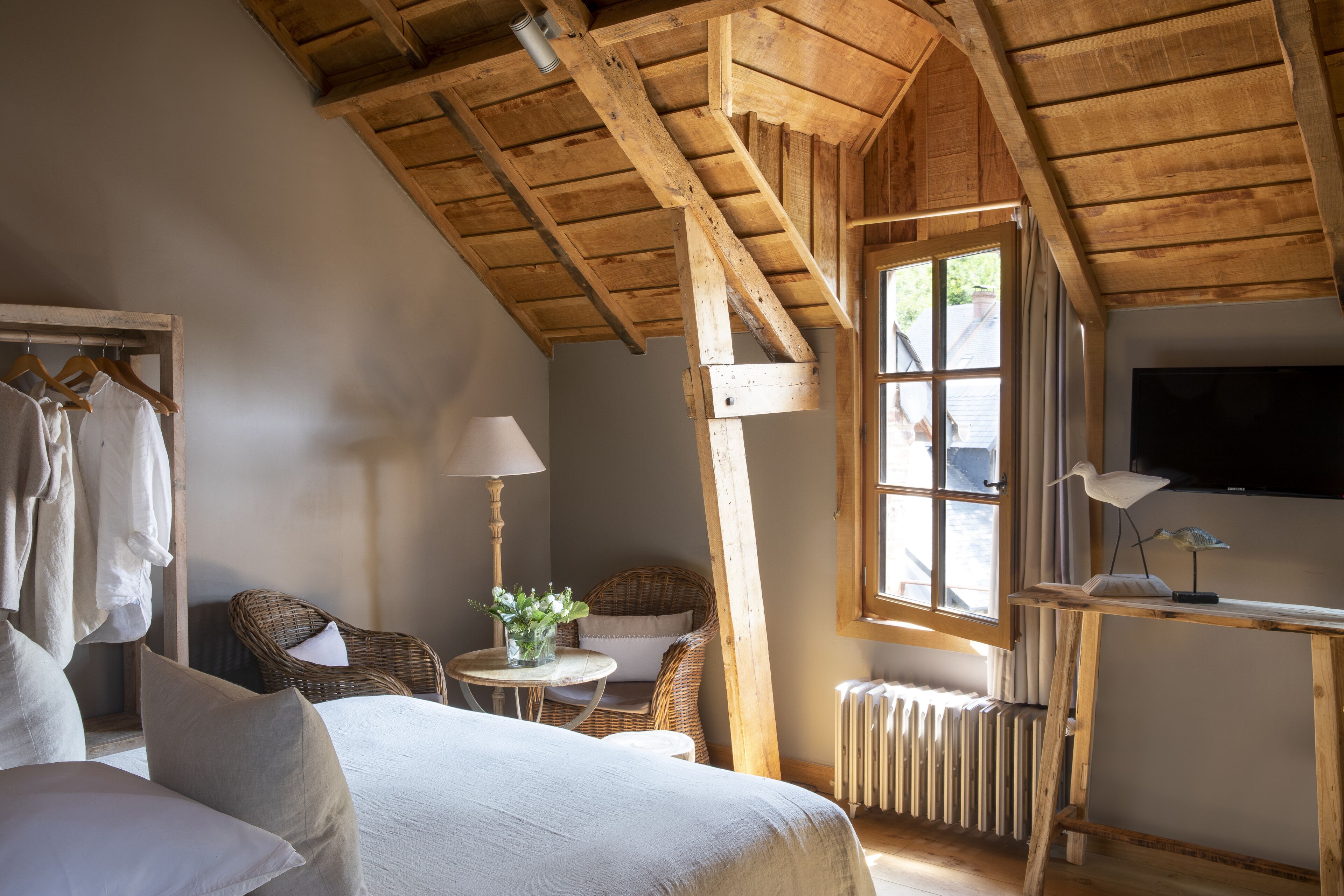 Una habitación acogedora con un alto techo de madera, una cama con sábanas blancas, una silla de mimbre, una pequeña mesa redonda con una lámpara y un televisor sobre un mueble de madera.