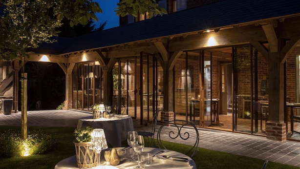 Un espacio de comedor al aire libre con mesas preparadas para dos, bajo un porche cubierto con paredes de ladrillo y puertas de cristal que dan a un espacio interior.