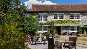 A stone building with a tiled roof, outdoor seating with wicker chairs and tables, and a green awning over the entrance.
