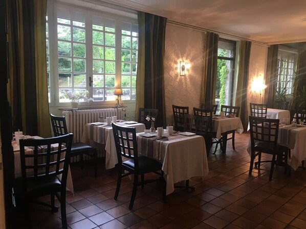 A dining area with multiple tables and chairs, large windows with curtains, and a radiator under one window.
