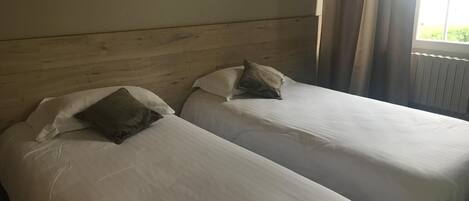 Two single beds with white linens and pillows, wooden headboard, and a window with curtains.
