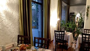 A dining area with a table set for breakfast, including bread, cheese, and ham. There are chairs around the table, and a potted plant is visible in the background.