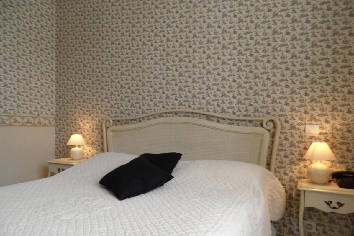 A neatly made bed with a white quilt and a black pillow, two bedside lamps, and a floral wallpaper pattern.