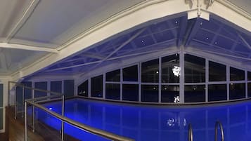 A modern indoor swimming pool with a glass ceiling, surrounded by seating areas and a staircase.
