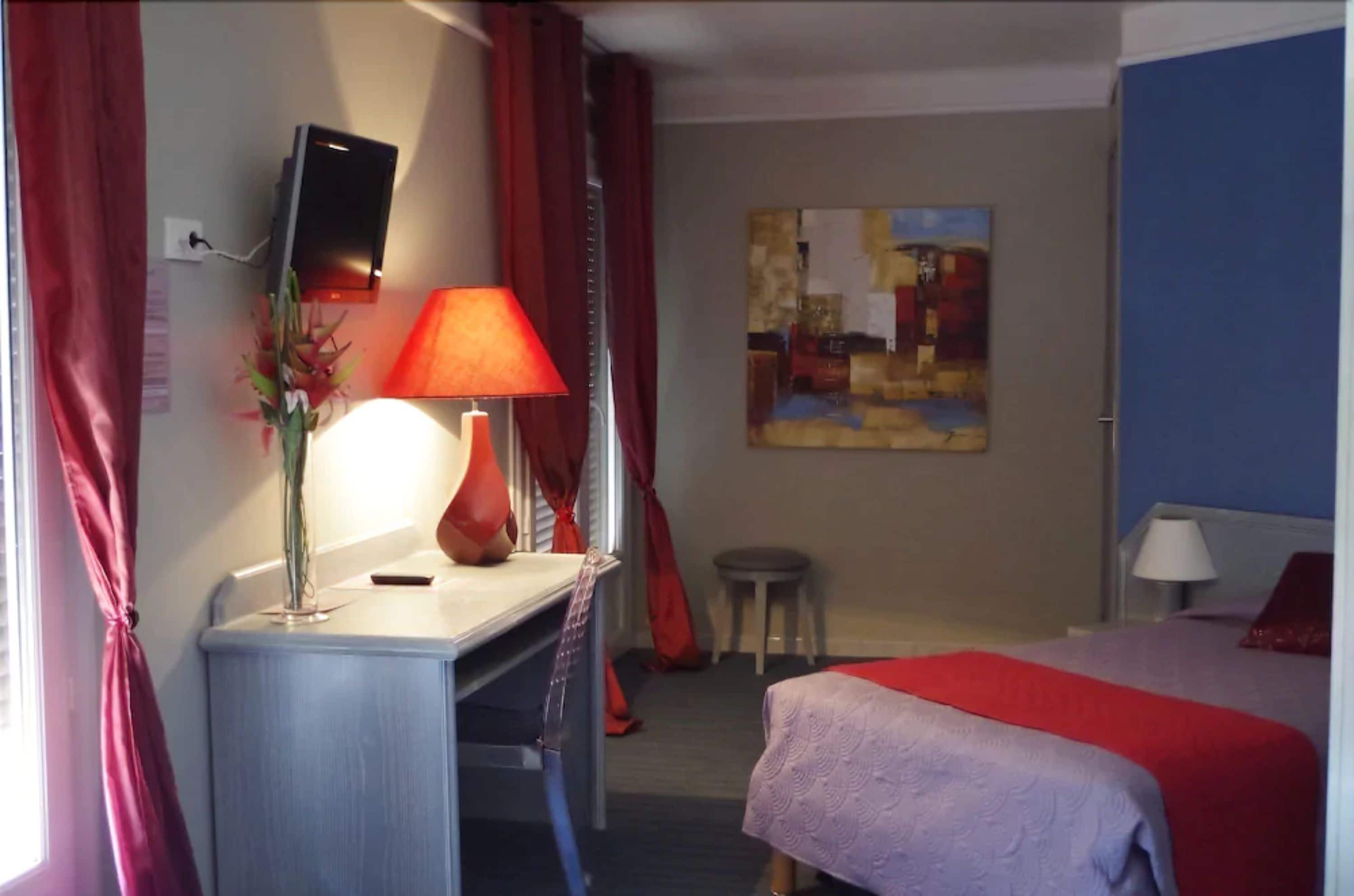 Habitación de hotel con escritorio, cama, televisor montado en la pared, un cuadro y ventana con cortinas rojas.