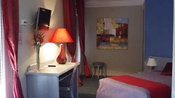 Habitación de hotel con escritorio, cama, televisor montado en la pared, un cuadro y ventana con cortinas rojas.