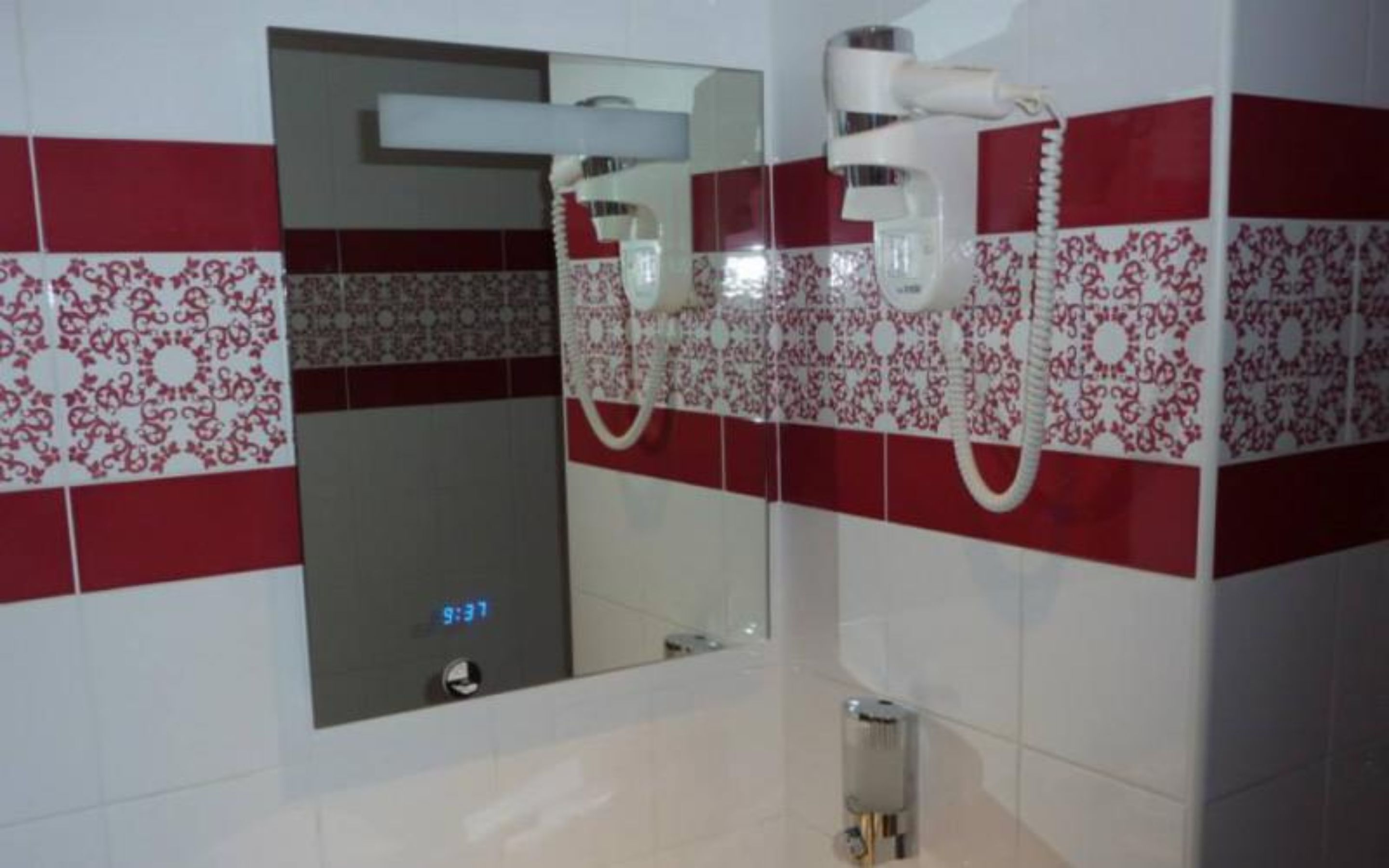 A bathroom with a mirror, red and white patterned tiles, a hairdryer, and a digital clock displaying 9:37.