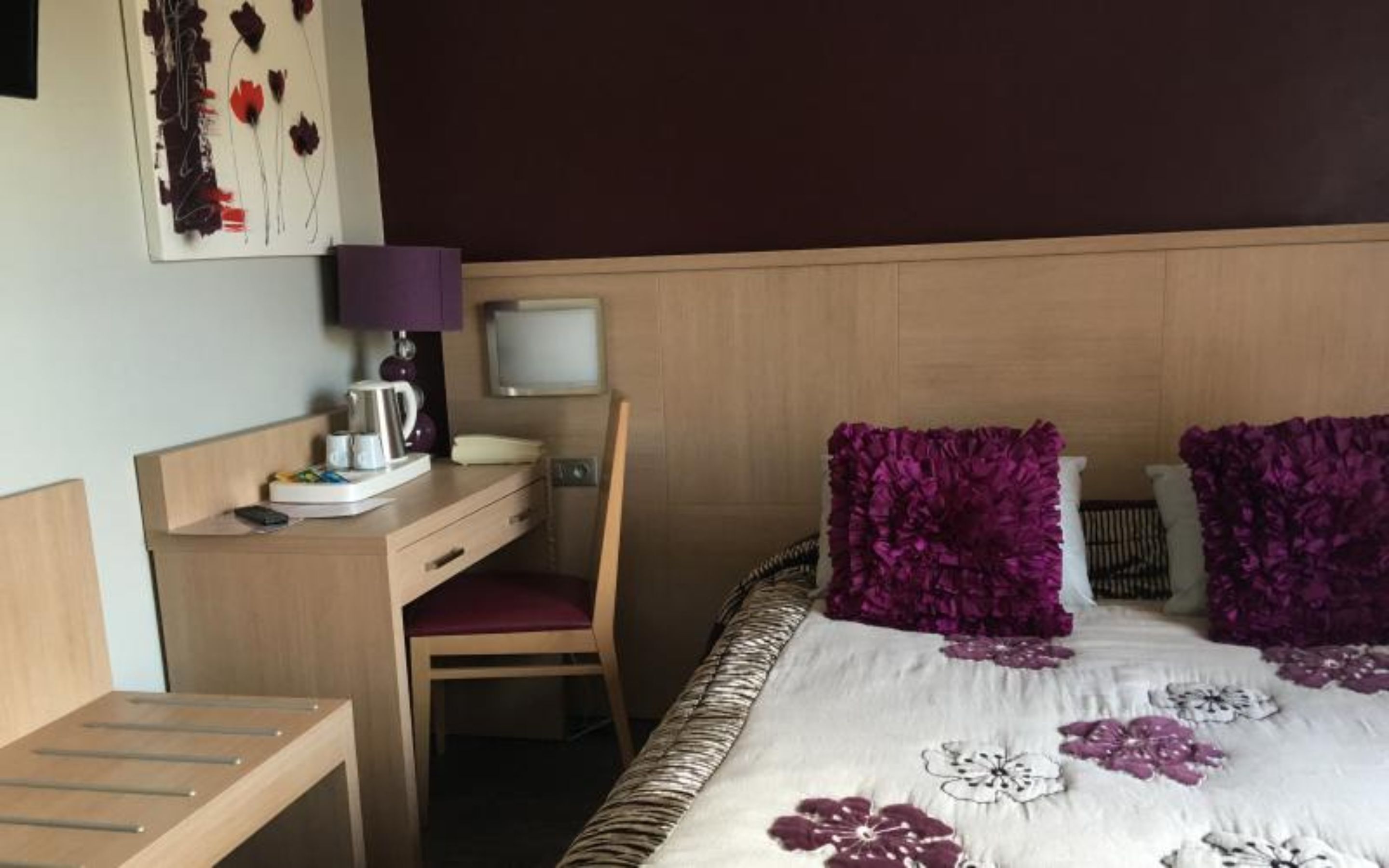 Una camera d'albergo con una testiera in legno, una scrivania con sedia, un letto con cuscini viola e una TV a muro.