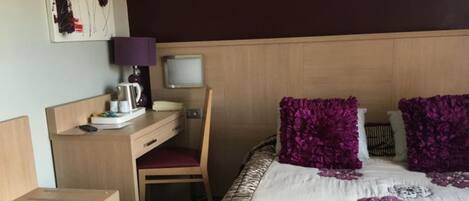 Habitación de hotel con cabecera de madera, un escritorio con silla, cama con almohadas moradas y televisor montado en la pared.