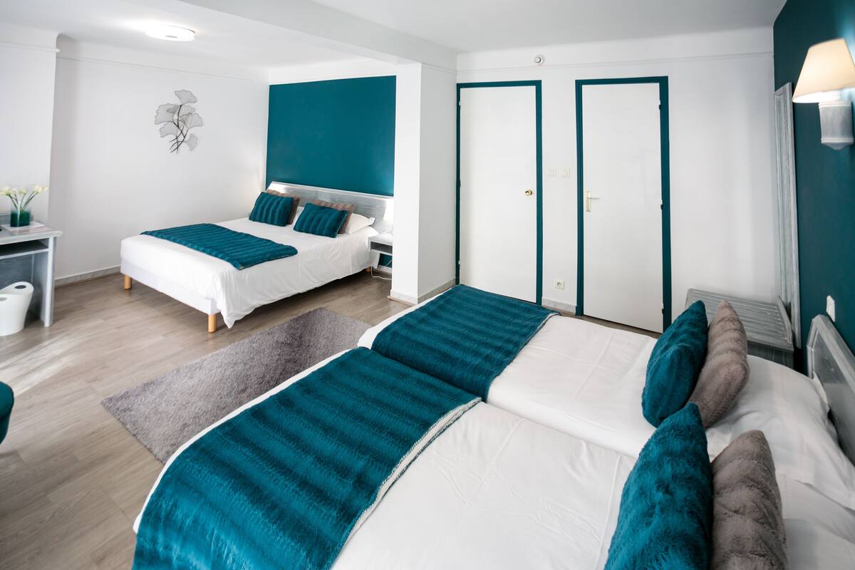 Ein modernes Schlafzimmer mit einem großen Bett, einem kleineren Bett und einem Schreibtisch mit Stuhl. Zwei Türen und eine Wandlampe sind vorhanden.