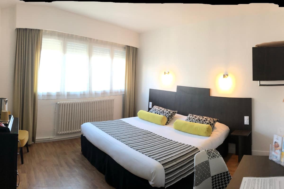 Habitación de hotel con cama, mesitas de noche, un escritorio con silla, radiador, ventana con cortinas y luces en la pared.