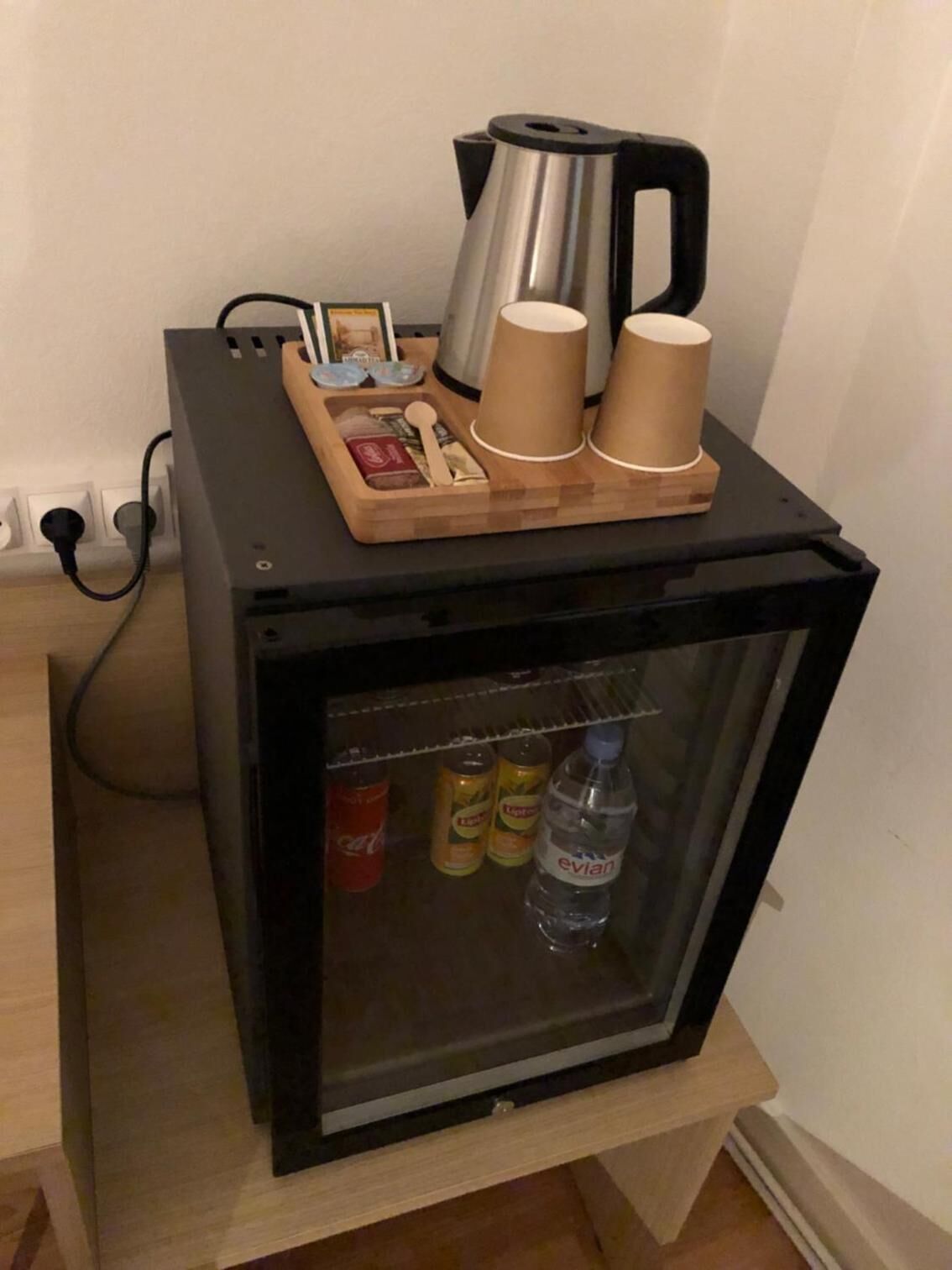 Un petit réfrigérateur noir, surmonté d’un plateau en bois contenant une machine à café, des tasses et une petite boîte. À l’intérieur du réfrigérateur, on trouve des boissons et une bouteille d’eau.