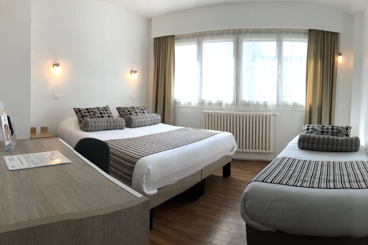 Una habitación de hotel con cama, escritorio, silla, una mesita y ventana con cortinas.