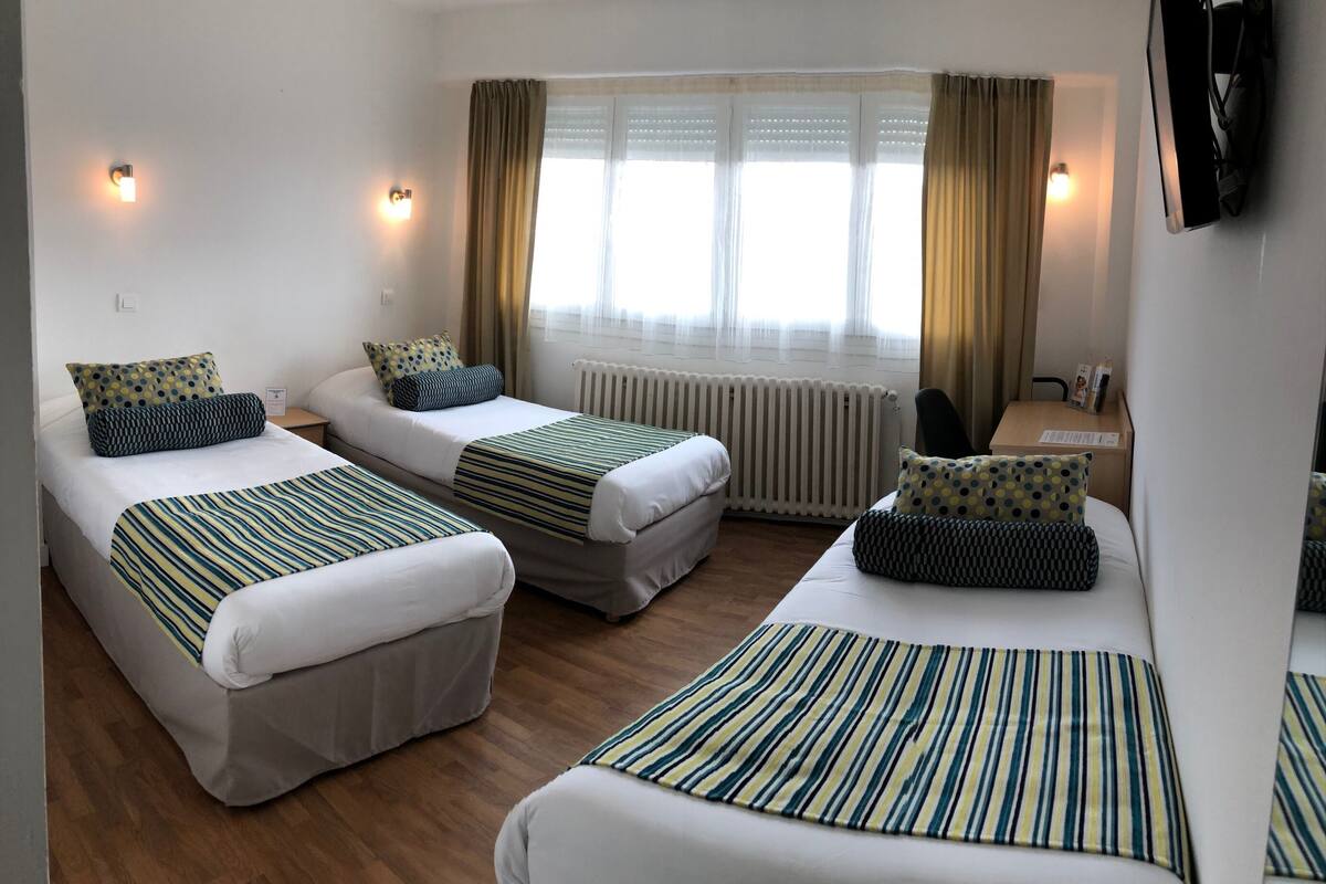 Habitación de hotel con dos camas individuales, una cama grande, televisor, ventana con cortinas y radiador.