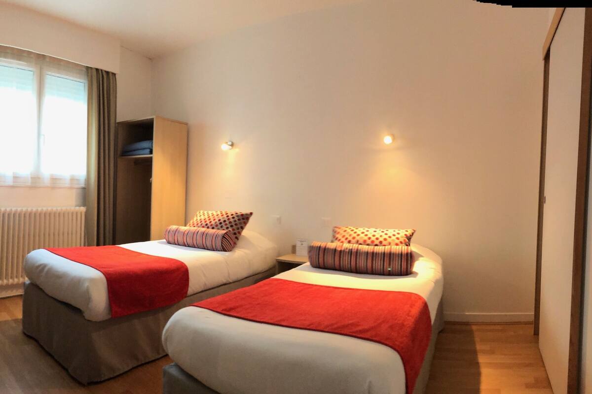 Habitación de hotel con dos camas, un escritorio, una silla, un radiador y una ventana con cortinas.