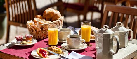 A breakfast table with croissants, pastries, a basket of bread, a coffee pot, cups, and a jug of orange juice.