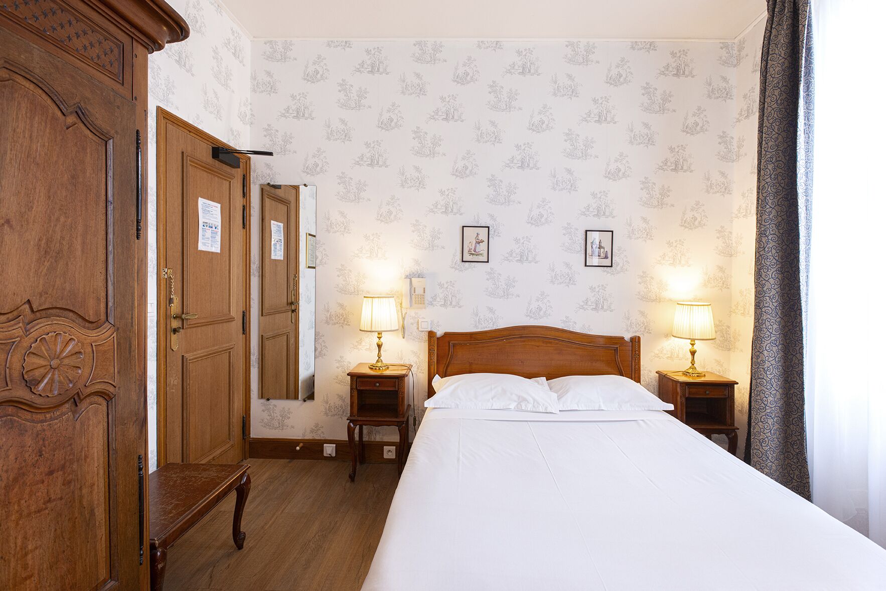 Habitación de hotel con armario de madera, cama con ropa blanca, dos mesitas de noche con lámparas y papel tapiz floral.