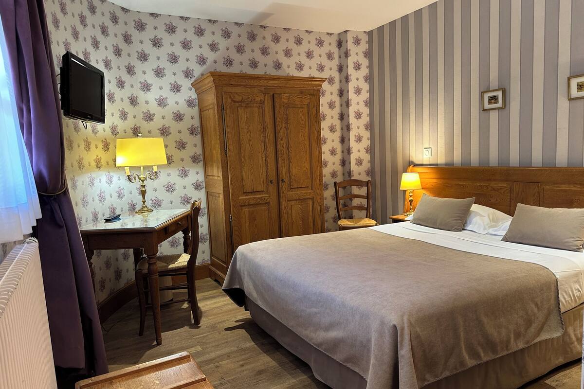 A hotel room with a bed, a wooden wardrobe, a desk with a lamp, and a television mounted on the wall.