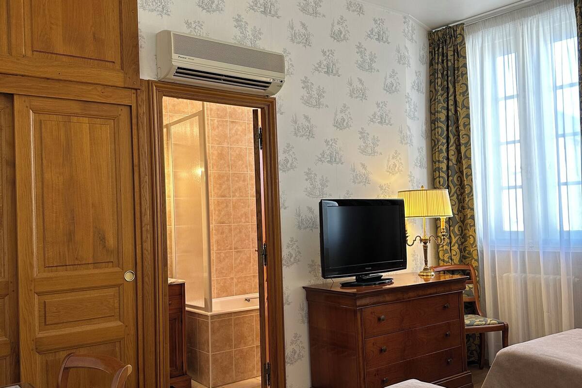 A hotel room with a wooden wardrobe, a television on a wooden cabinet, two beds with beige bedding, a chair, and a window with curtains.