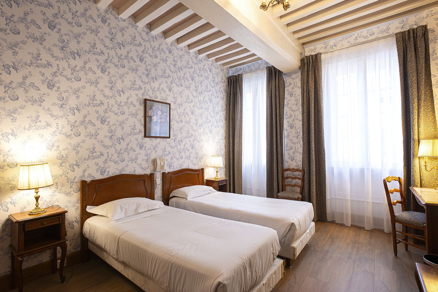 A hotel room with two beds, wooden headboards, floral wallpaper, a wooden ceiling, a desk with a chair, and a telephone on the wall.