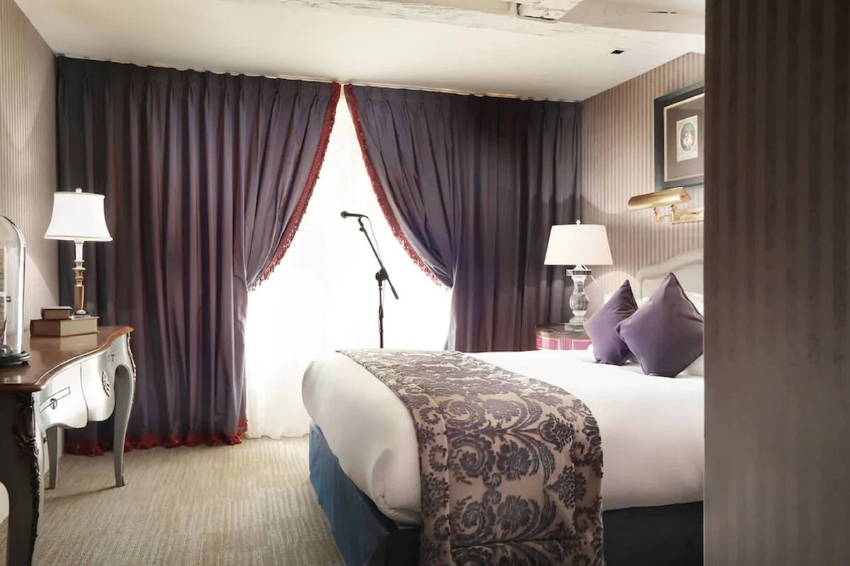 A hotel room with a bed, two bedside lamps, a desk with a lamp, a microphone stand, and a framed picture on the wall.