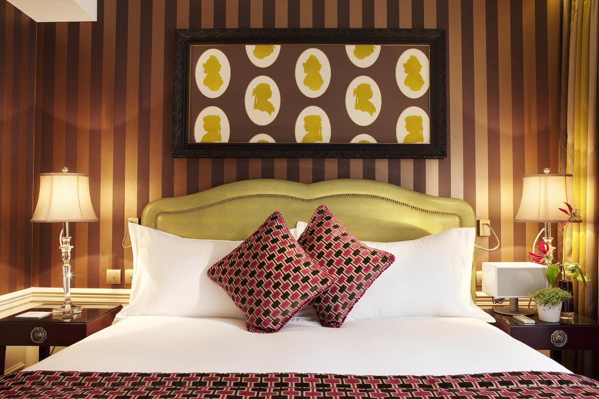 A hotel room with a large bed, two nightstands, a headboard, a framed picture, and a potted plant.