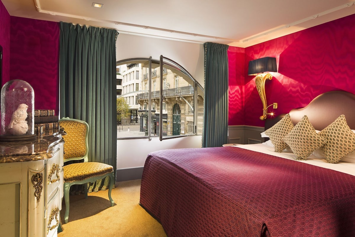 A hotel room with a bed, a chair, a dresser, a lamp, and a view of the city through a window.