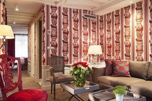 Une chambre d’hôtel avec un papier peint à motifs, un canapé, un fauteuil, une table ornée d’un vase de fleurs et une lampe.