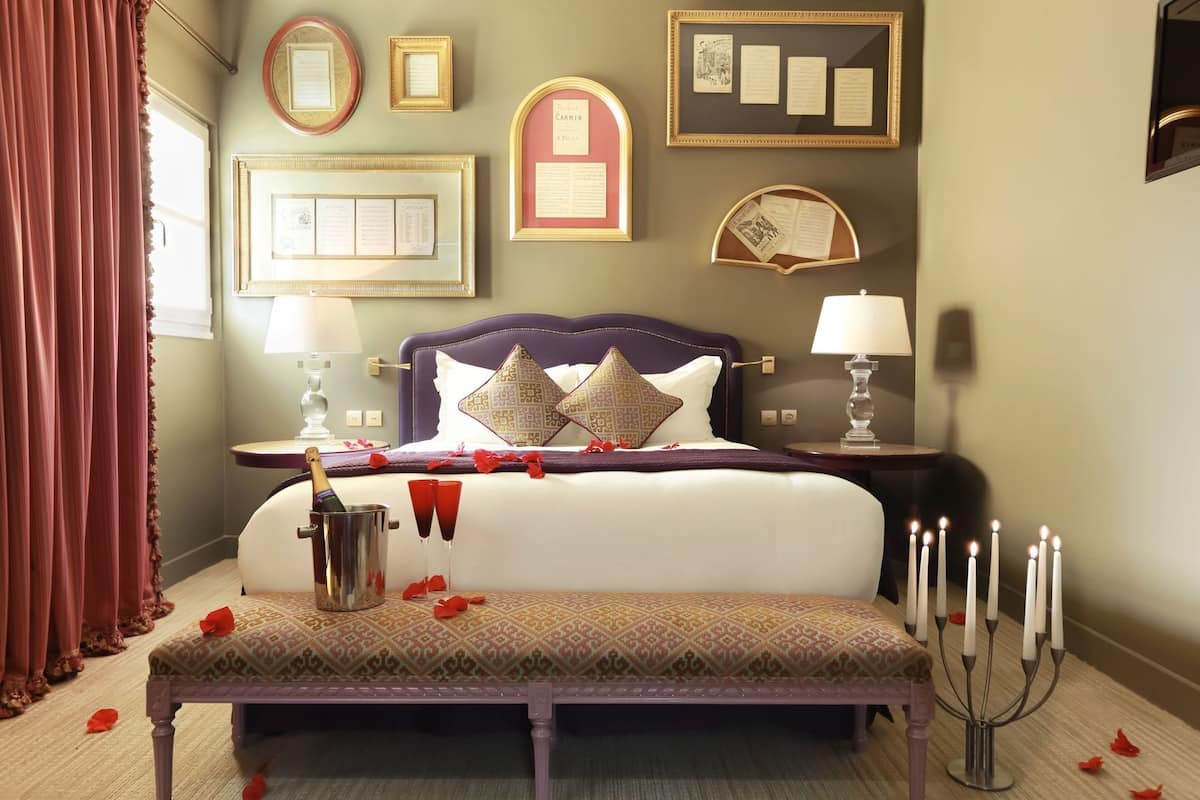A bedroom with a bed, bedside tables, a bench, a wall with framed items, and a window with curtains.